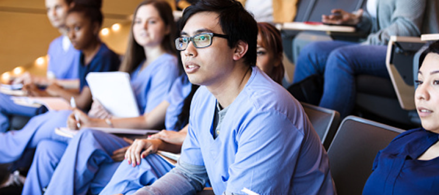 Medical school enrollment hits new record. Med school students in scrubs sitting in a lecture hall listening to a professor.